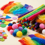 Craft kit supplies showing fuzzy rainbow pipe cleaners, colorful pom-poms, feathers, foam shapes and pastel buttons on a cream background.