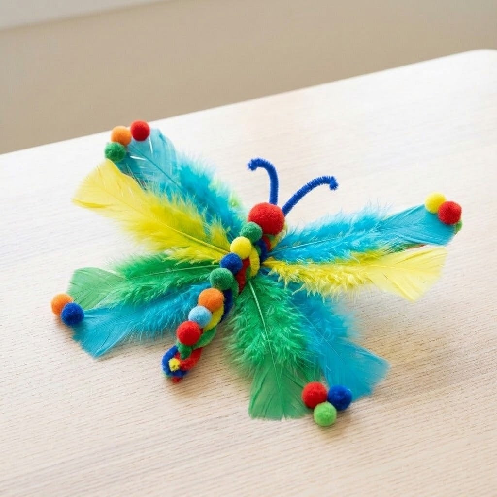 Craft kit project example showing a butterfly with blue, yellow and green feather wings, colorful pom-poms and pipe cleaners on pale wood.