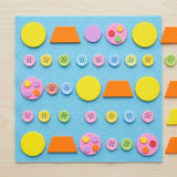 A colorful children's sensory craft kit on a light blue felt board, featuring yellow circles, orange trapezoids, pink and purple circles, and multicolored buttons, displayed on a light wood background.
