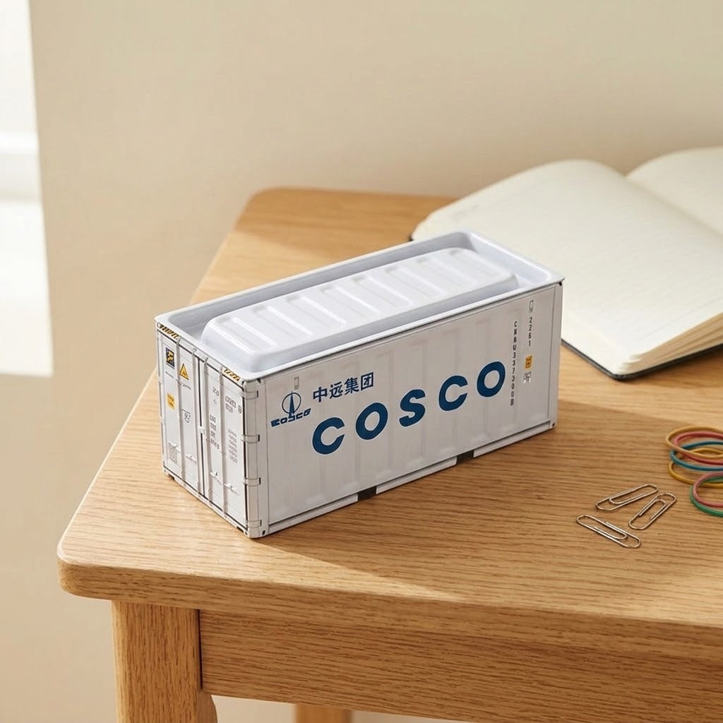 A white iron storage box with blue lettering sits on a wooden desk beside an open notebook, paper clips, and rubber bands.