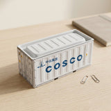 A white storage box with a recessed ribbed lid and blue lettering sits on a light wooden desk beside two paper clips.