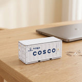 A white storage box with its lid closed sits on a wooden desk beside paper clips and a closed silver laptop.