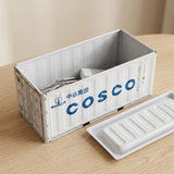 A white shipping container-style storage box with blue COSCO branding sits on a light wood table, with its matching lid placed beside it.