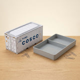 A white storage box stands beside a gray two-compartment tray and scattered paper clips on a wooden desktop.