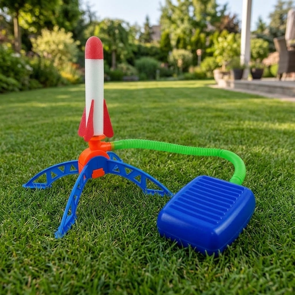 A plastic toy rocket launcher set on green grass in a backyard. The set includes a red, white, and orange rocket, a blue support stand, a green hose, and a blue foot pedal.