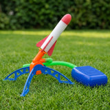 A plastic toy rocket launcher set on green grass outdoors, featuring a white and red rocket, blue and orange launch stand, green hose, and a blue rectangular control or accessory piece.