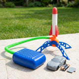 Rocket launcher with blue foot pump and red-and-white rocket on outdoor paving beside a toy car, loose batteries, and a gray cover.