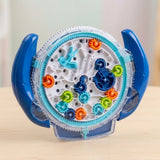 A blue-handled transparent plastic maze puzzle with colourful gears stands on a light wooden tabletop against a softly blurred background.