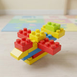 A colorful toy building block structure made of interlocking red, yellow, green, blue, and beige blocks, standing on a light wood surface with a blurred pastel play mat in the background.