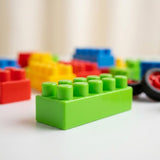 A bright green toy building block with studs, shown in sharp focus in the foreground with other colorful blocks blurred in the background on a white surface.