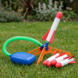 A rocket launcher stands on grass with an orange base, blue pedal, green hose, and red-and-white foam rockets.