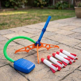 A toy rocket launcher set on a stone patio with grass in the background. The set includes an orange and blue launcher stand, a blue foot pedal connected by a green hose, and several white and red foam rockets lined up in front.