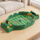 Green tabletop soccer game with yellow controls and a ball at midfield on a wooden coffee table beside a cream sofa.