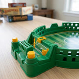 Close-up of a green tabletop soccer game's yellow buttons, goal, and numbered sliding score rail, with board games behind.