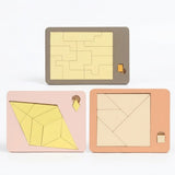 Three wooden geometric puzzle designs show interlocking, diamond and square arrangements in grey, pink and peach trays against a white background.