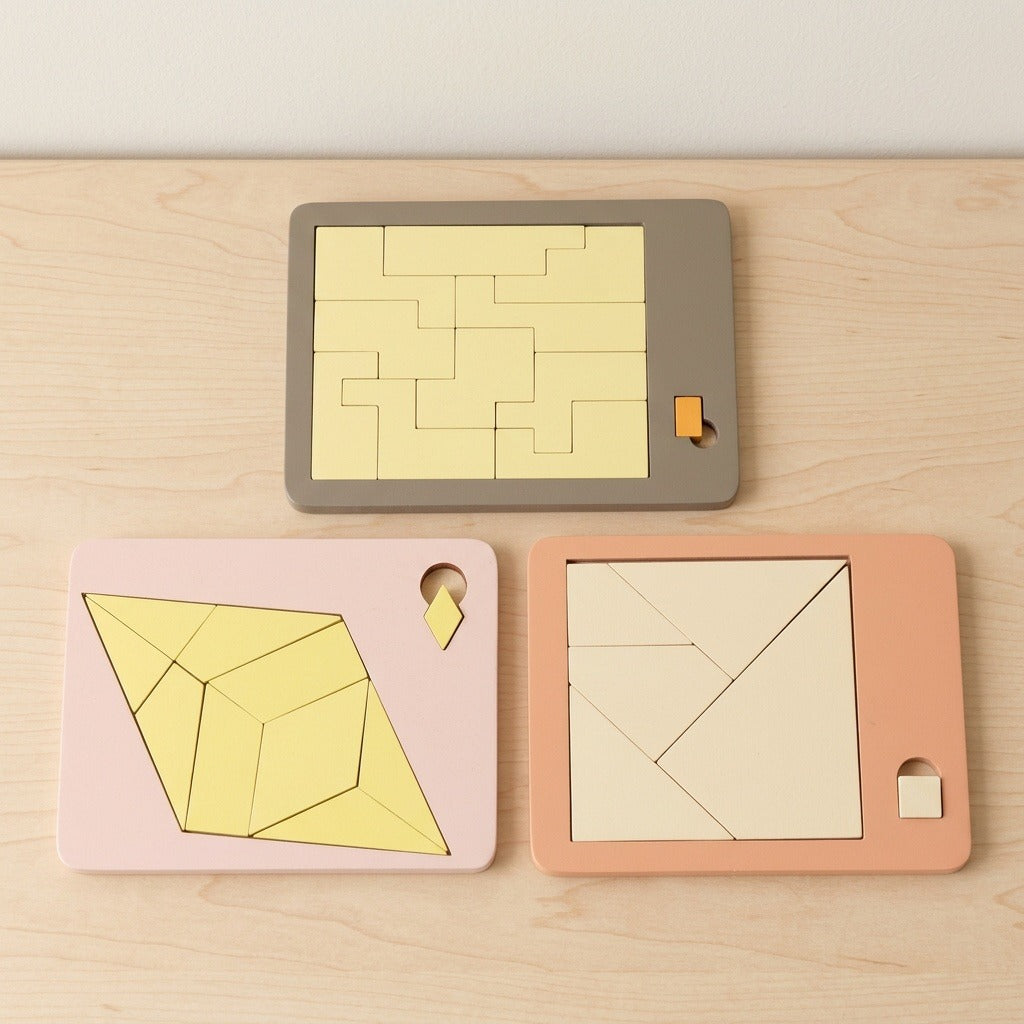 Three geometric puzzle boards in gray, pink, and peach frames with yellow and cream pieces rest on a light wooden table.
