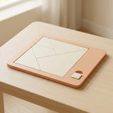 Cream tangram pieces fitted into a peach geometric puzzle board on a light wooden table beside a window.