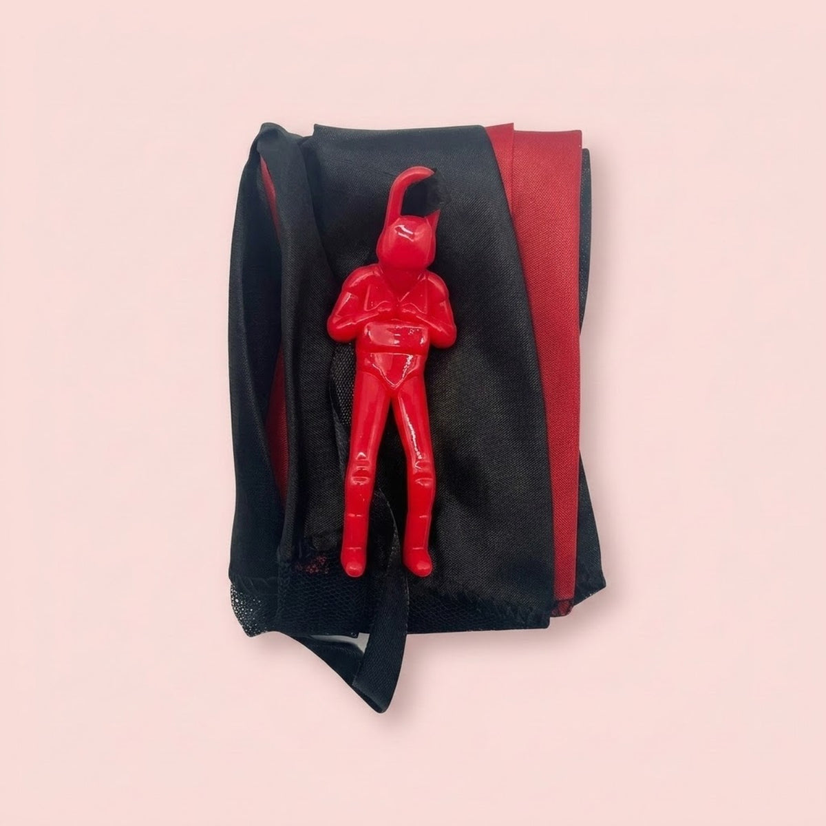 Folded red and black parachute toy with its red plastic soldier figure on top, photographed against a plain pale pink background.
