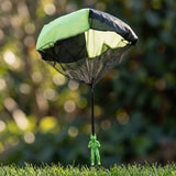 Green and black toy parachute with a mesh skirt hovering above green grass, its green plastic soldier hanging from a single black strap.