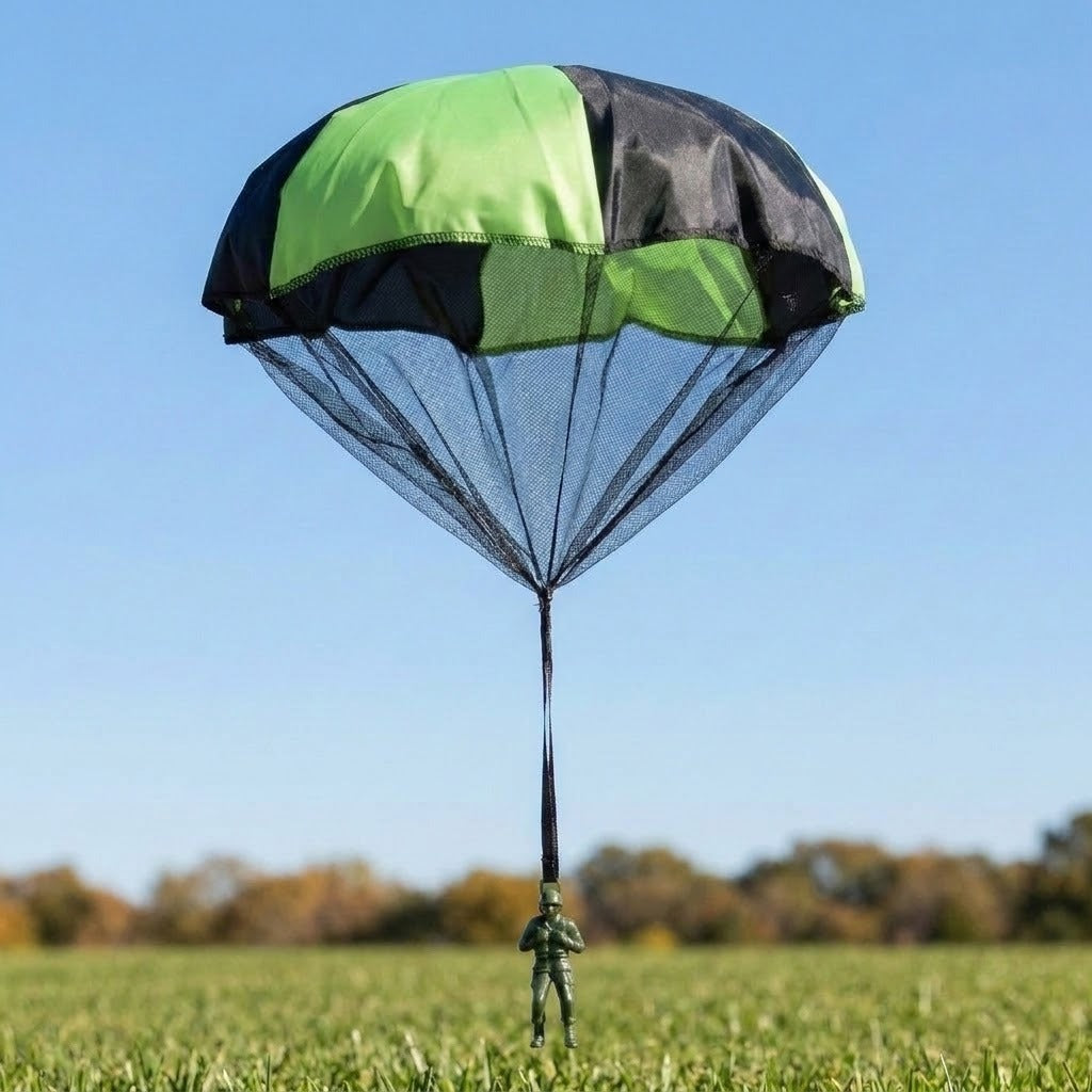 Green and black toy parachute with a mesh skirt and single strap holding a dark green plastic soldier, over a grass field under blue sky.