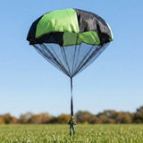 Green and black toy parachute with a mesh skirt and single strap holding a dark green plastic soldier, over a grass field under blue sky.