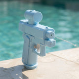 Light blue water squirt gun sprays a narrow stream from its front nozzle while standing on wet poolside paving.