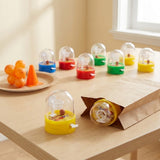 Colorful mini basketball games sit on a wooden table beside orange decorations and an open brown paper bag containing another game.