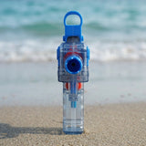 A blue and transparent water gun stands facing forward on sand with blurred turquoise waves behind it.