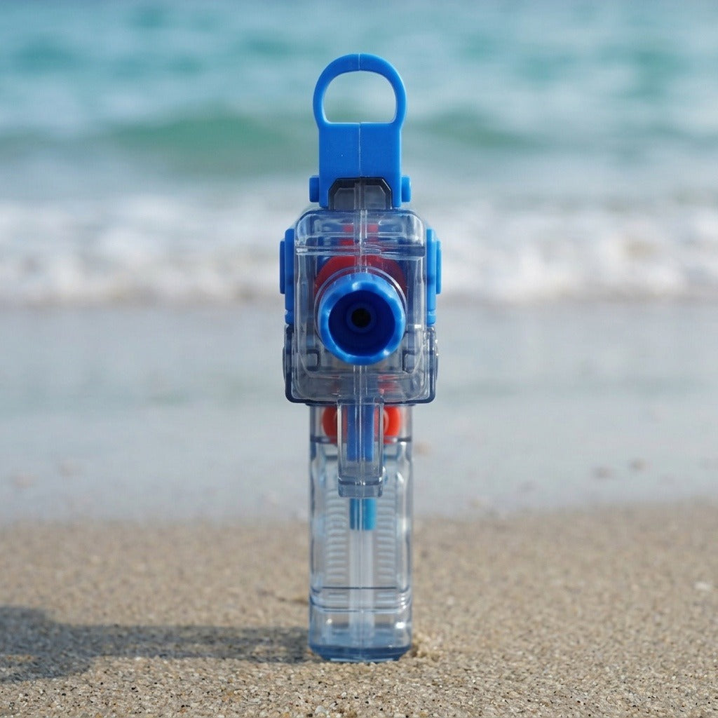 A blue and transparent water gun stands facing forward on sand with blurred turquoise waves behind it.