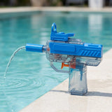 A blue and transparent water gun with an orange trigger stands on a pool edge, squirting a curved stream into the water.