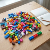 Multicolored interlocking building blocks lie scattered on a wooden table beside an open notebook, pencil, and sharpener.