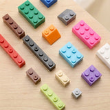 A set of colorful toy building block pieces arranged on a light wood surface. The pieces come in multiple colors including pink, green, blue, orange, purple, brown, gray, red, and white, with different stud counts.