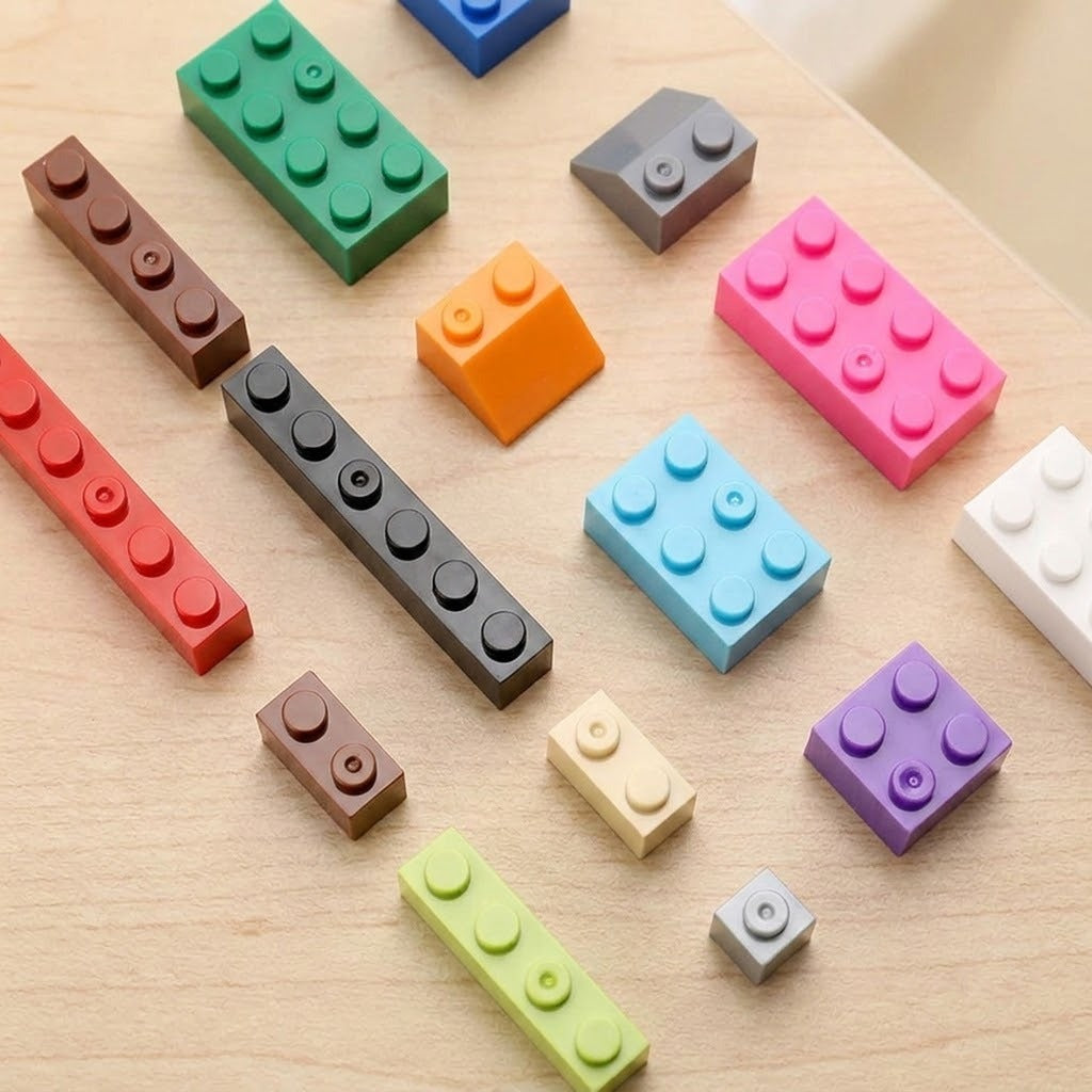 A set of colorful toy building block pieces arranged on a light wood surface. The pieces come in multiple colors including pink, green, blue, orange, purple, brown, gray, red, and white, with different stud counts.