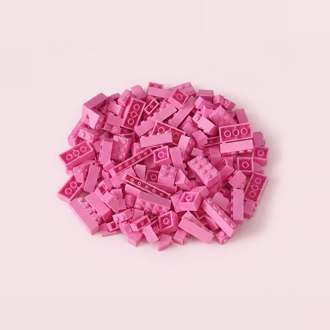 Pink plastic building blocks in assorted rectangular sizes form a compact pile on a pale pink background.