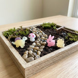A miniature fairy garden arrangement in a square wooden tray, featuring small figurines including a yellow turtle, pink-and-white mushroom, pink bunny, and yellow frog, set in soil with pebbles, moss, and a small twig on a light wood table.