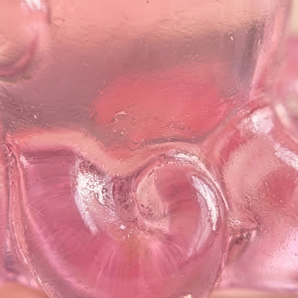 A close-up of a translucent pink resin miniature figurine reveals glossy curled details and tiny surface marks.