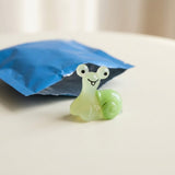 A pale green resin miniature snail figurine with a smiling face sits beside an opened blue blind bag on a cream surface.