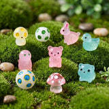 Pink and blue resin miniature animal figurines, spotted mushrooms and round ornaments rest on bright green moss among scattered pebbles.