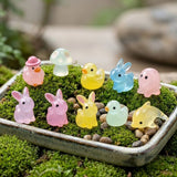 Ten pastel resin miniature figurines shaped like animals and a mushroom sit among moss and pebbles in a shallow rectangular tray.