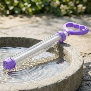 A purple and clear plastic water gun rests across a water-filled stone basin outdoors, with its nozzle touching the water.