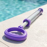 A purple pull-out water gun with a clear barrel lies on a light stone deck beside a swimming pool.