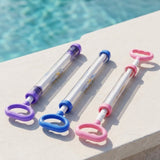 Three pull-out water guns with clear bodies and purple, blue, and pink handles lying on a white stone ledge beside a swimming pool.