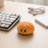 A small orange-brown cute squishy toy with a smiling face sits on a light wood desk next to a keyboard, USB cable, and white notebook.
