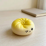 A small yellow and cream squishy toy shaped like a round bun or dumpling with a simple black face, sitting on a light wood tabletop with a blurred book in the background.
