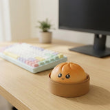 An orange steamed-bun squishy toy sits in a brown round container on a wooden desk beside a pastel keyboard and monitor.
