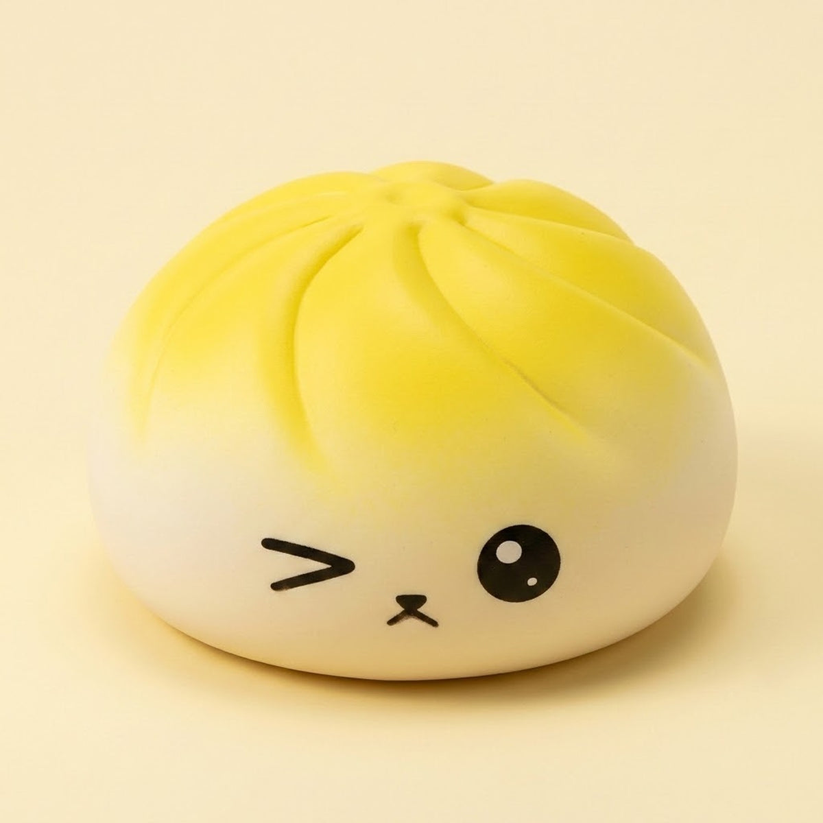 A cute, winking dumpling-shaped squishy toy with a yellow-to-white gradient and black facial features, set against a solid pale beige background.