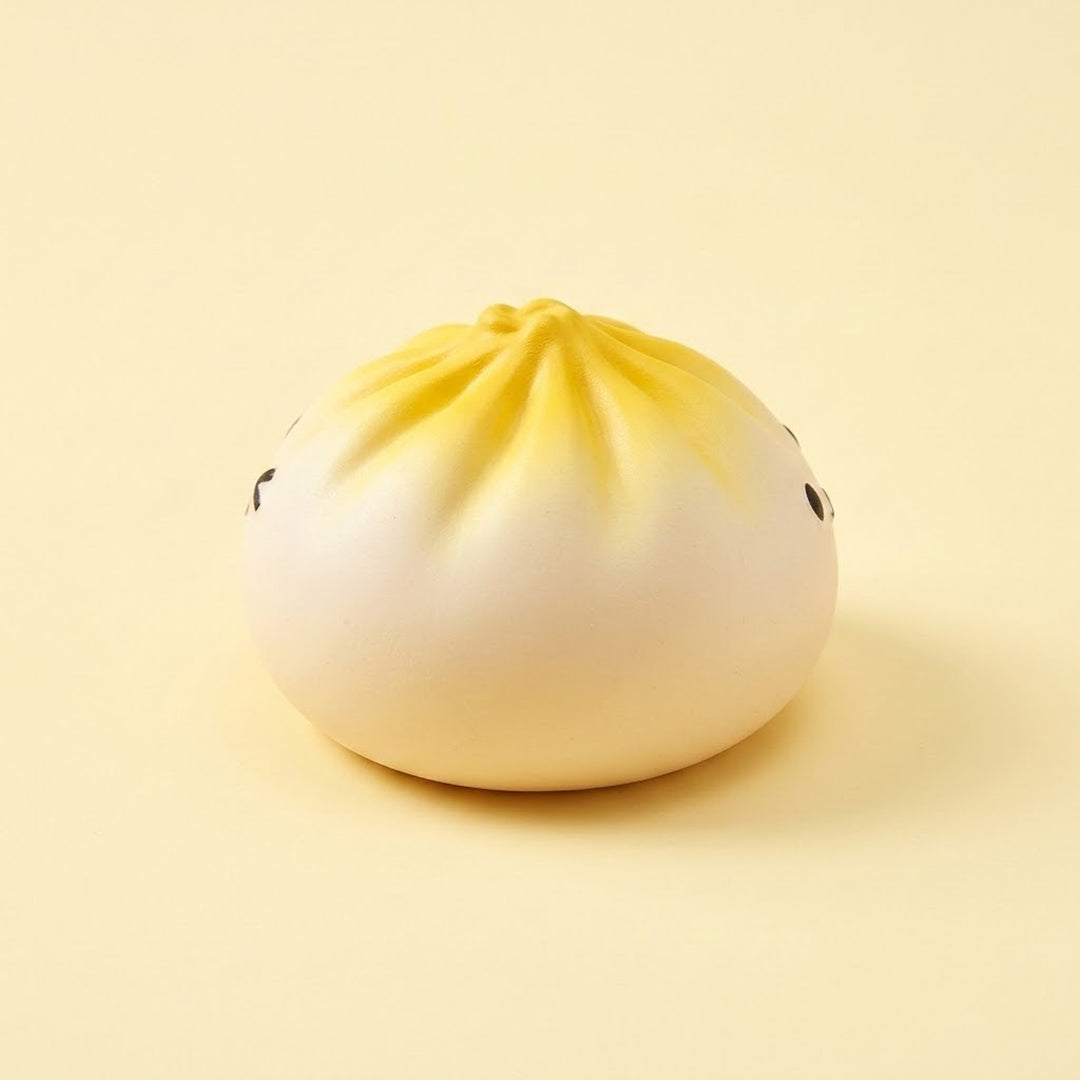 Round white squishy toy with a yellow pleated top and small black eyes rests on a pale yellow surface against a matching background.