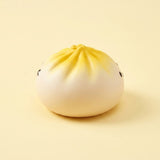 Round white squishy toy with a yellow pleated top and small black eyes rests on a pale yellow surface against a matching background.