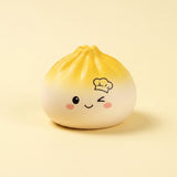 A cute, round, dumpling-shaped squishy toy with a yellow-to-white gradient, featuring a winking face, pink cheeks, and a small chef hat illustration on the forehead, set against a solid pale yellow background.