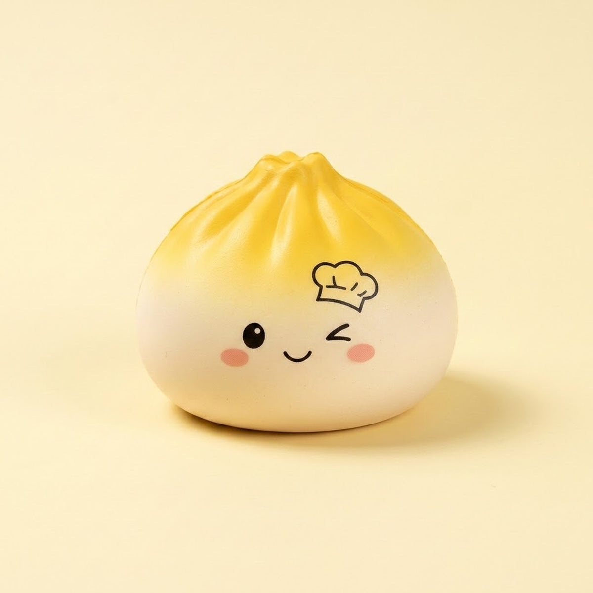 A cute, round, dumpling-shaped squishy toy with a yellow-to-white gradient, featuring a winking face, pink cheeks, and a small chef hat illustration on the forehead, set against a solid pale yellow background.